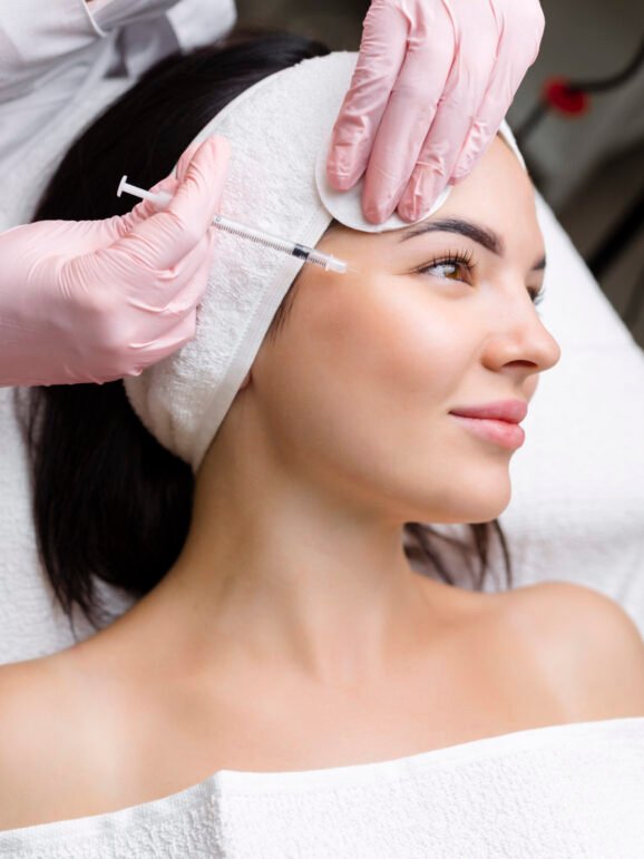 Close up of hands of young cosmetologist injecting botox in female face. She is standing and smiling. The woman is closed her eyes with relaxation Close up of hands of young cosmetologist injecting botox in female face. She is standing and smiling. The woman is closed her eyes with relaxation.