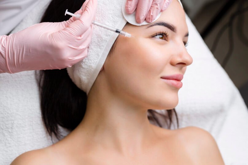 Close up of hands of young cosmetologist injecting botox in female face. She is standing and smiling. The woman is closed her eyes with relaxation.