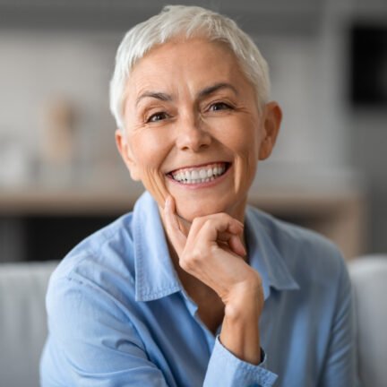 Portrait of lovely older gray woman smiling to camera and touching chin, sitting on sofa at home indoors. Happy beautiful mature lady resting and relaxing in living room, expressing happiness
